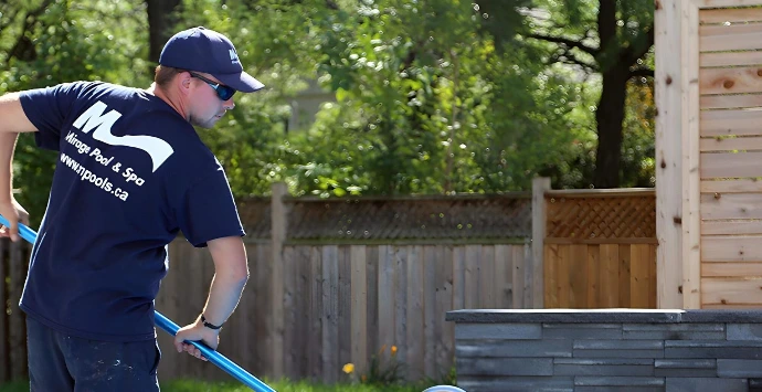 A person cleans a swimming pool with a skimmer in a backyard setting, surrounded by trees and a wooden fence.