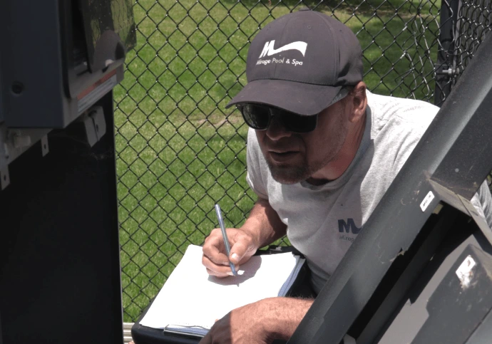 A person kneels by a large gas heater, taking notes on a clipboard, with a fenced grassy area in the background.